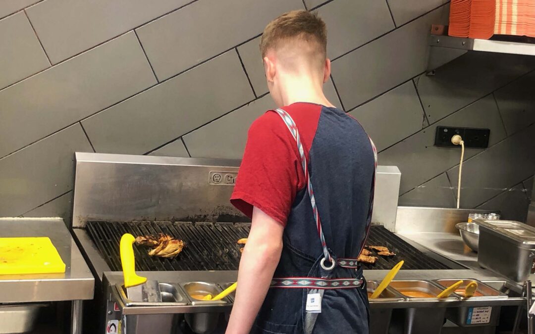 Young person wearing an apron and working as a chef, standing at a grill cooking chicken.