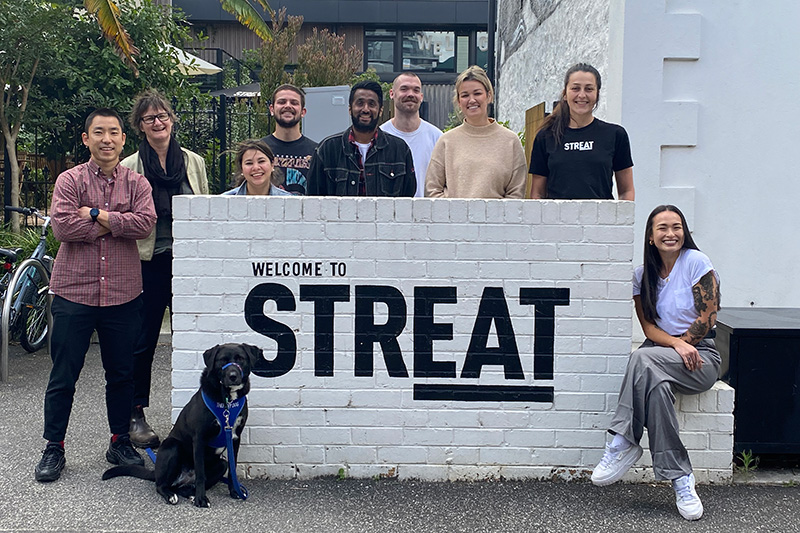 Nine STREAT youth programs staff and therapy dog smiling outside the STREAT cafe sign.