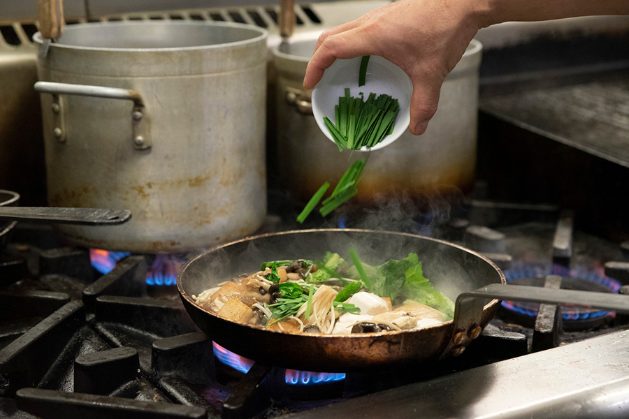 A chef's hand is tipping chives into a pan of mushrooms, cooking on a gas stove.