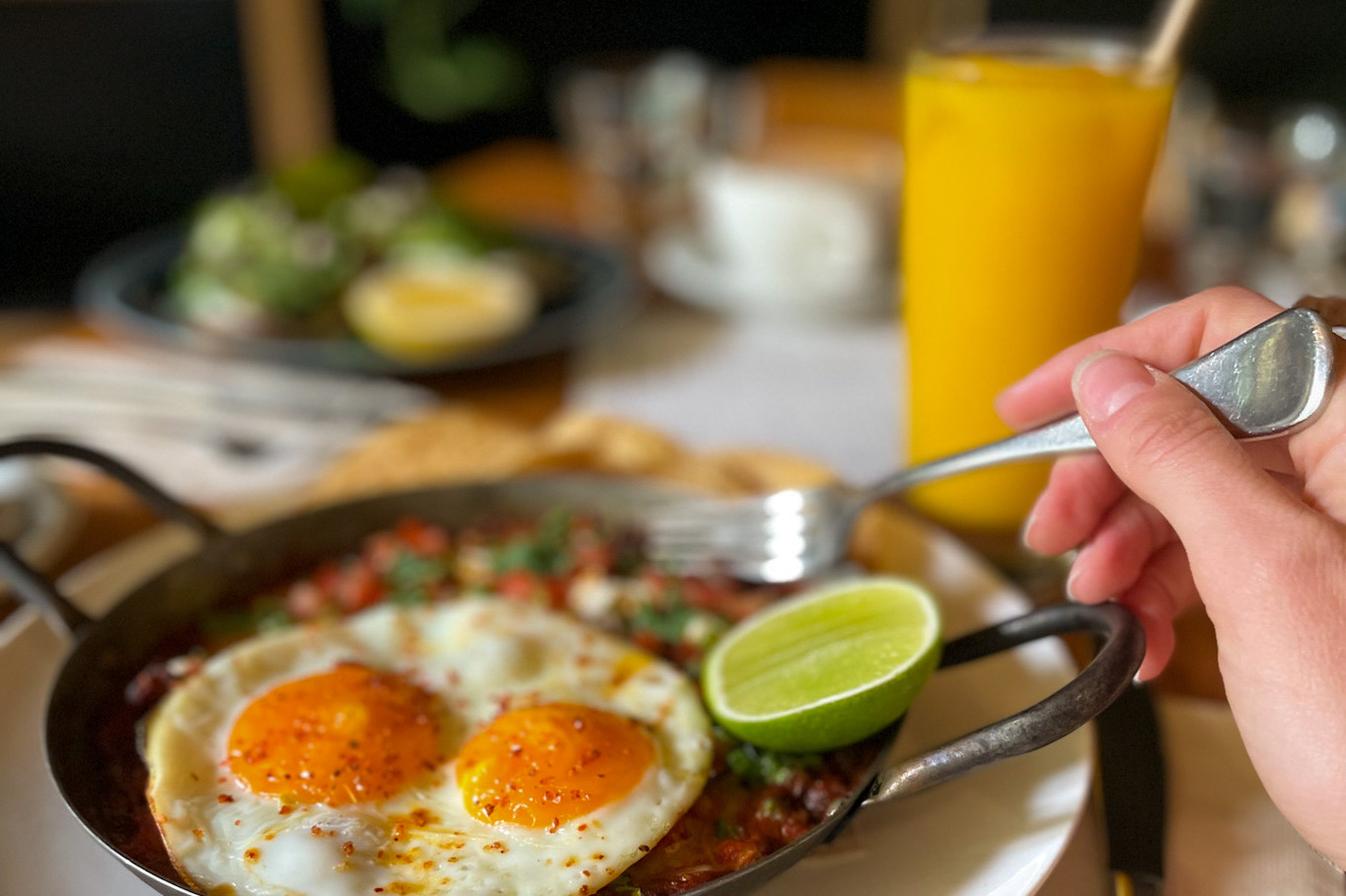 A skillet of Mexican-style baked eggs with a lime slice, two fried eggs, on a cafe table with plants in the background and a glass of fresh orange juice.