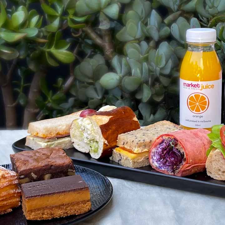 Selection of STREAT Catering on a table: Freshly-made sandwiches, wraps, caramel slice, chocolate brownie, a chia pudding with fruit, fresh orange juice and STREAT coffee on a green table cloth in front of some plants.