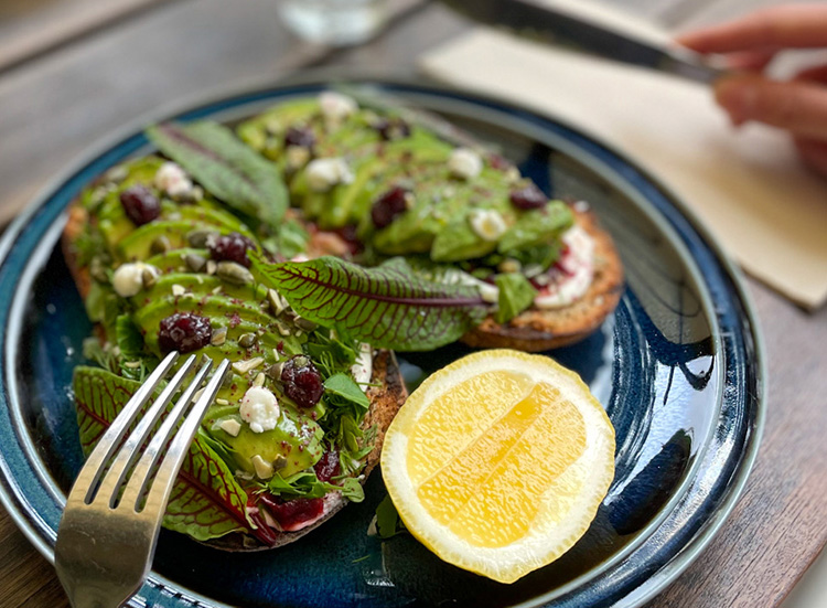 A close up of a blue plate with avocado on toast, a lemon slice, red sorrel, feta and olive tapenade.