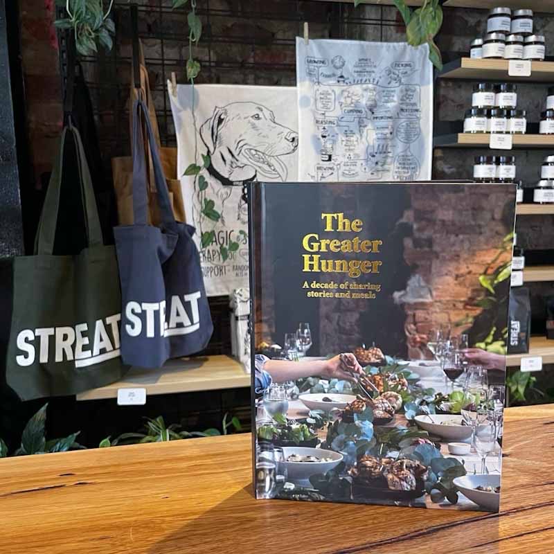STREAT's cookbook, The Greater Hunger, standing on a table with STREAT tote bags and tea towels in the background.