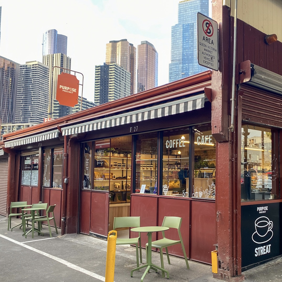 External shot of STREAT's Cafe at Vic Market, showing Melbourne CBD high rise buildings behind, and a maroon sign for the Purpose Precinct.